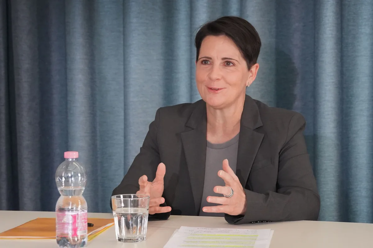 Eine Frau mit kurzen schwarzen Haaren sitzt hinter einem Tisch, vor ihr ein Wasserglas und Dokumente.